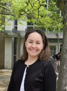 Veronika smiling. She has shoulder-length brown hair, is wearing a black cardigan over a white shirt, stands in front of a building with green trees in the background.